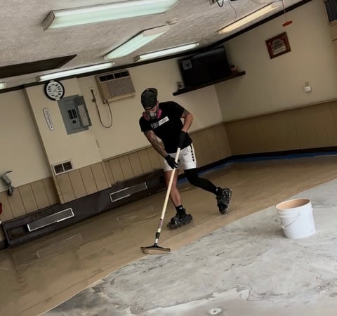 Person mopping wet floor in empty gymnasium with concrete surface and fluorescent lighting